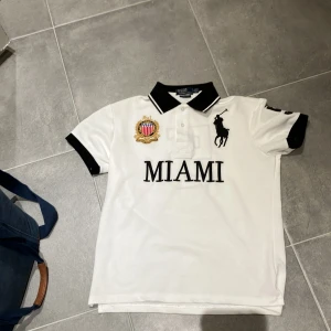 Vit pikétröja från Ralph Lauren - Säljer en snygg vit pikétröja från Ralph Lauren med svart krage och ärmslut. Tröjan har broderad logga och texten 'MIAMI' på framsidan samt siffran '5' på ryggen. Perfekt för en stilren look!