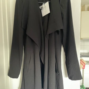 Svart trenchcoat från H&M - Snygg svart trenchcoat från H&M med knappar och bälte i midjan. Jackan har långa ärmar och en stilren design som passar perfekt för höst och vår. Perfekt för att hålla stilen i alla väder!