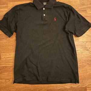 Svart pikétröja från Ralph Lauren - Säljer en klassisk svart pikétröja från Ralph Lauren med den ikoniska röda broderade logotypen på bröstet.           Tröjan är i ganska fint skick.           Priset är ej hugget i sten!