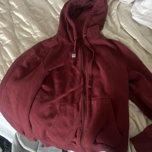 Vinröd hoodie från Sinsay - Säljer en vinröd hoodie från Sinsay med dragkedja och huva. Tröjan har långa ärmar och en praktisk ficka framtill. Perfekt för en avslappnad stil.