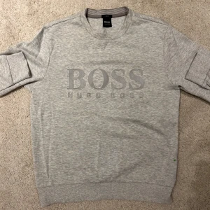 Hugo Boss tröja S - Snygg grå sweatshirt från Hugo Boss med stort BOSS-tryck på framsidan. Tröjan har långa ärmar och en rund halsringning. Storlek S. Hugo Boss