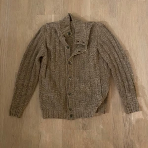 Cardigan från Ferrante - Säljer en snygg beige stickad kofta från Ferrante. Koftan har långa ärmar och knappar framtill. Perfekt för kyligare dagar och ger en stilren look. Passar bra till jeans och sneakers.