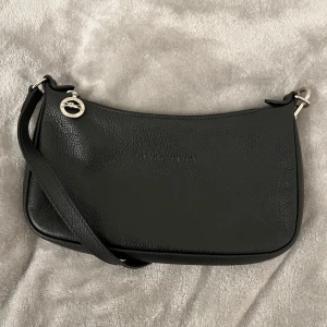 Svart axelväska från Longchamp - Snygg svart axelväska från Longchamp i skinn. Väskan har en elegant design med en diskret logotyp präglad på framsidan och en dragkedja för säker förvaring. Perfekt för att bära dina nödvändigheter med stil.