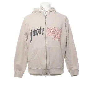 Beige hoodie från Wasted - Säljer en beige hoodie från Wasted med tryck i svart och rött framtill. Hoodien har dragkedja och en klassisk känguruficka. Perfekt för en avslappnad stil.