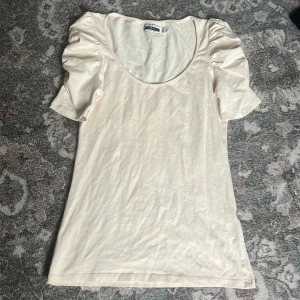 Beige topp från Zara - Säljer en söt beige topp från Zara med korta puffärmar och rund halsringning. Toppen är i ett mjukt material och har en enkel design som passar perfekt till vardags. Perfekt för dig som vill ha något stilrent och bekvämt.