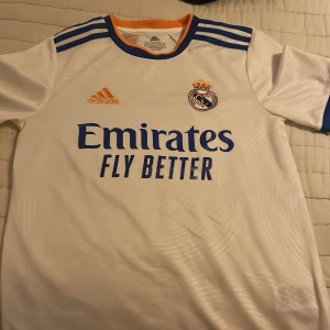Vit Real Madrid fotbollströja från Adidas - Säljer en vit Real Madrid fotbollströja från Adidas med korta ärmar. Tröjan har blå detaljer och klubbens emblem på bröstet. Den är tillverkad i Primegreen-material och har en orange och blå krage. Perfekt för alla fotbollsfans! Är även äkta därför säljer jag för ett dyrt pris. 🙃 pruta gärna 