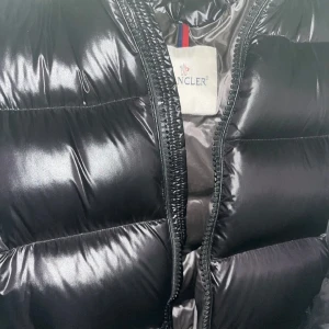 Svart pufferjacka från Moncler - Snygg svart pufferjacka från Moncler med glansigt material och dragkedja. Jackan har en klassisk design med Moncler-logga på armen. Perfekt för kyliga dagar.