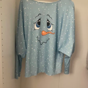 Blå långärmad tröja med snöflingor och ansikte från Sheilay - Säljer en blå långärmad tröja från Sheilay med ett lekfullt ansikte och snöflingor som mönster. Perfekt för en avslappnad och rolig stil. Tröjan har en lös passform och är tillverkad i ett mjukt material.
