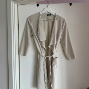Beige cardigan från Boohoo - Säljer en stilren beige kappa från Boohoo. Den har en elegant design med knytband i midjan och långa ärmar. Perfekt för att ge en chic touch till din outfit.
