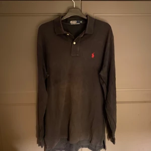 Svart pikétröja från Ralph Lauren - Säljer en klassisk svart pikétröja från Ralph Lauren med den ikoniska loggan broderad i rött på bröstet. Tröjan har långa ärmar och en stilren krage med knappar. Perfekt för en avslappnad men ändå stilfull look.