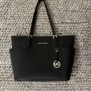 Svart Michael Kors väska - Snygg svart väska från Michael Kors med silverfärgade detaljer och logotyp. Väskan har en praktisk dragkedja och flera fack inuti för enkel organisering. Perfekt för att bära med sig det viktigaste med stil.
