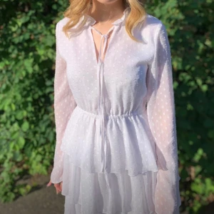 Student klänning - Charmig vit klänning med prickigt mönster och volangdetaljer. Den har långa ärmar och en v-ringad hals med knytband. Perfekt för en somrig look!