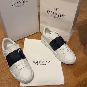 Valentino Garavani sneakers med svart band - Snygga vita Valentino Garavani sneakers med ett brett svart band över snörningen. Skorna har en stilren design med subtila nitar på hälen och är tillverkade i skinn. Perfekta för en trendig look. Skorna är knappt använda mycket bra skick!