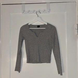 Grå ribbad tröja från Gina tricot - Säljer en stilren grå ribbad tröja från Gina. Tröjan har en v-ringad hals och är croppad med långa ärmar. Perfekt för en avslappnad look.