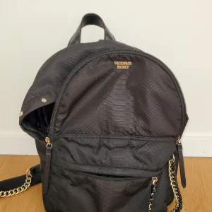 Svart ryggsäck från Victoria's Secret - Snygg svart ryggsäck från Victoria's Secret med ormskinnsmönster. Väskan har guldfärgade kedjedetaljer och flera praktiska fack med dragkedja. Perfekt för att bära dina saker med stil.