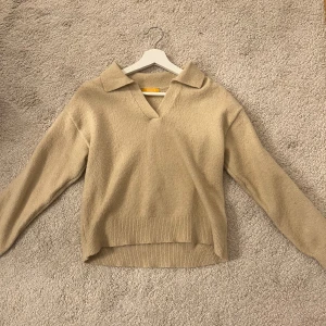 Beige stickad tröja med krage - Säljer en stilren beige stickad tröja med krage och v-ringning. Tröjan har långa ärmar och ribbade kanter vid ärmslut och nederkant. Perfekt för kyligare dagar!