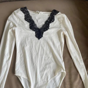 Vit långärmad body från H&M - Säljer en stilren vit body från H&M med långärmad design och vacker svart spetsdetalj vid halsringningen. Perfekt för att lägga till en touch av elegans i din garderob. Passar bra till både jeans och kjol.