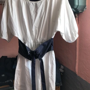 Vit klänning med marinblå detaljer - Snygg vit klänning med marinblå bälte och detaljer. Klänningen har korta ärmar och en elastisk midja för en bekväm passform. Perfekt för en stilren look.