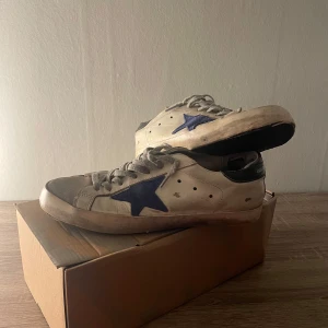 Golden Goose sneakers med blå stjärna - Säljer ett par Golden Goose sneakers med en distinkt blå stjärna på sidan. Skorna har en vit bas med svarta detaljer vid hälen och snörning framtill. Perfekta för en avslappnad och stilren look. Orginal Box medföljer vid köp. Defekter syns på bild 3-5 därför är det det låga priset. Tror att defelterna går att fixa hos sko makare. Priset kan diskuteras vid snabbt köp 