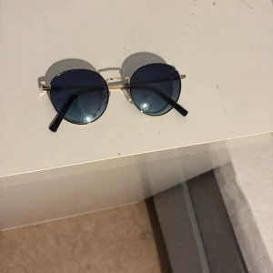 Runda solglasögon med blå linser - Snygga runda solglasögon med blåtonade linser och guldfärgade bågar. De har svarta skalmar som ger en stilren look. Perfekta för att ge din outfit en cool touch.