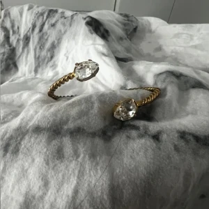 Caroline svedbom armband 🥰 - Elegant guldarmband med en tvinnad design och två stora, klara kristaller i ändarna. Perfekt för att ge en glittrig touch till din outfit.