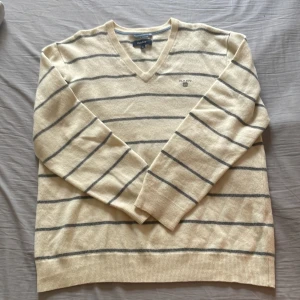 Beige randig tröja från Gant - Säljer en stilren beige tröja från Gant med grå ränder och v-ringning. Tröjan är tillverkad i 100% lammull och har långa ärmar. Perfekt för en avslappnad och elegant look. Står xl men passar bäst s/m. Köpt sacond hand men nyskick