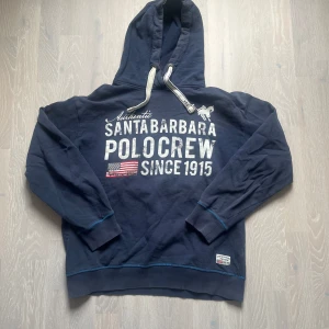 Polo hoodie - Säljer en mörkblå hoodie med tryck från Santa Barbara Polo & Racquet Club. Tröjan har en stor text på framsidan och en amerikansk flagga. Den har en justerbar huva med vita snören och ribbade muddar. Riktig fet hoodie med en liten defekt på under armen! 