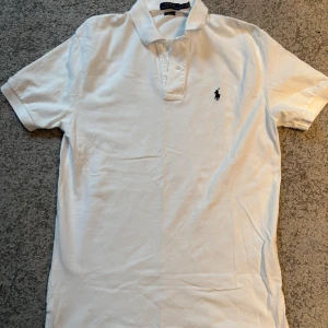 Vit pikétröja från Ralph Lauren - Säljer en klassisk vit pikétröja från Ralph Lauren med korta ärmar och broderad logga på bröstet. Perfekt för en stilren och avslappnad look. Storlek L.