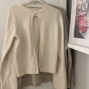 Beige kofta med knappdetalj - Säljer en stilren beige kofta med rund hals och en enkel knapp framtill. Koftan har långa ärmar och ribbade kanter som ger en klassisk look. Perfekt för lager-på-lager under kyligare dagar.