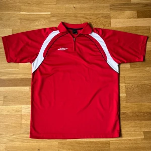 Vintage Umbro Röd T-Shirt/Fotbollströja - Vintage Umbro T-Shirt/Fotbollströja i färgen röd med piké krage och dragkedja. Storlek: L, Armhåla till Armhåla: 57 cm, Längd: 72.5 cm, Krage till Armöppning: 41 cm.  Material: 100% Polyester.