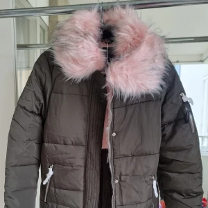 Olivgrön pufferjacka med rosa fuskpäls - Säljer en superfin olivgrön pufferjacka med en mysig rosa fuskpäls runt kragen. Jackan har långa ärmar och dragkedja framtill. Den är perfekt för kalla vinterdagar och har praktiska fickor med dragkedjor. Insidan är fodrad med mjukt rosa tyg för extra värme och komfort. Passar perfekt för både vardag och festliga tillfällen!