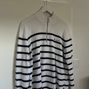 Vit och svart randig tröja med dragkedja - Säljer en snygg vit tröja med mörkblå ränder från Bondelid. Den har en halv dragkedja framtill och en hög krage. Perfekt för en stilren och avslappnad look. Passar bra till både vardag och fest!