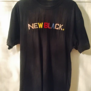 Svart t-shirt från New Black - Säljer en svart t-shirt från New Black med färgglad text på bröstet. T-shirten är i storlek L och har en klassisk passform med korta ärmar. Perfekt för en avslappnad stil! Trådarna på texten har börjat släppa lite.
