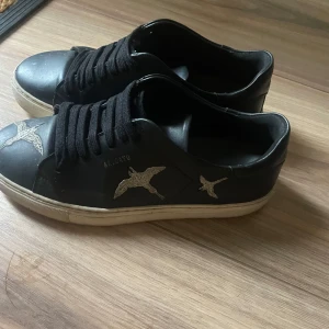 Arigatos - Säljer ett par svarta sneakers med coola fågelmotiv på sidorna. Skorna har en vit sula och svarta snören. Perfekta för en avslappnad stil och passar till det mesta i garderoben.