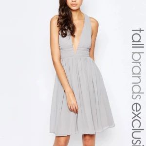 Grå klänning med djup urringning - Säljer en elegant grå klänning med djup urringning och korsade axelband i ryggen. Klänningen har en vacker, flödande kjol och är perfekt för festliga tillfällen. Den är ärmlös och har en smickrande passform som framhäver midjan.