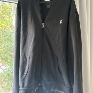 Svart hoodie med dragkedja från Ralph Lauren - Säljer en svart hoodie från Ralph Lauren med dragkedja och det klassiska logotypbroderiet på bröstet. Perfekt för en avslappnad stil! Köpt på Joels butik för 1799kr Inga defekter förutom att snöret inte finns kvar.