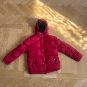 Röd dunjacka från Polo Ralph Lauren - Säljer en snygg röd dunjacka från Polo Ralph Lauren. Jackan är perfekt för vintern med sin varma vaddering och långa ärmar. Den har en praktisk huva och stängs med både dragkedja och knappar. Insidan är fodrad i en mörkblå färg som ger en fin kontras