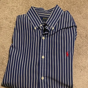 Blå randig skjorta från Ralph Lauren - Snygg blå skjorta från Ralph Lauren med vita ränder och klassisk krage. Skjortan har knappar framtill och en broderad röd logotyp på bröstet. Perfekt för en stilren look.