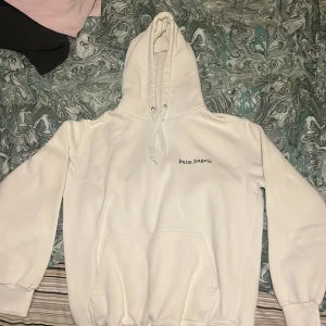 Vit hoodie från Palm Angels - Säljer en stilren vit hoodie från Palm Angels med deras ikoniska logga tryckt både fram och bak. Hoodien har en klassisk passform med långa ärmar och en praktisk ficka framtill. Perfekt för en avslappnad look.färgen bak har släppt lite men inte mätbart alltså på texten 