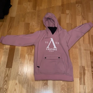 Rosa hoodie från Slade Couture - Säljer en snygg rosa hoodie från Slade Couture med tryck framtill och baktill. Den har en praktisk magficka med knappar och en bekväm huva. 