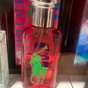 Röd parfymflaska från Ralph Lauren - Snygg parfymflaska i röd färg från Ralph Lauren med en grön logotyp av en polospelare. Flaskan har en elegant och stilren design med en silverfärgad kork. Perfekt för den som vill ha en doft med stil och attityd.