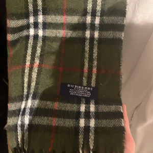 Grön rutig halsduk från Burberry - Säljer en snygg grön rutig halsduk från Burberry. Den har klassiska rutor i vitt, svart och rött. Gjord av mjuk ull och perfekt för att hålla dig varm under kyliga dagar. Ett stilrent val för både höst och vinter.