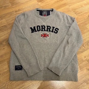 Morris tröja - Säljer en stilren grå tröja från Morris med broderad text och brittisk flagga på framsidan. Tröjan har långa ärmar och ribbade muddar vid ärmslut och nederkant. Perfekt för en avslappnad och snygg look.