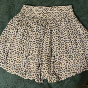 Blommig kortkjol med volang - Söt kortkjol med blått blommönster på vit bakgrund. Kjolen har en volangdetalj och en elastisk midja för bekväm passform. Perfekt för en somrig look! Finns också shorts under.