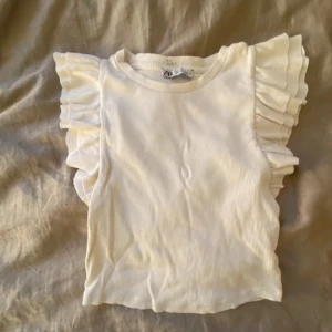 Topp med volang från zara - Säljer en söt vit ribbad topp från Zara med volangdetaljer på ärmarna. Perfekt för en stilren och romantisk look. Toppen är ärmlös och har en mjuk och bekväm passform.