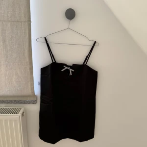 Svart axelbands klänning med rosett - Elegant svart klänning med smala axelband och en söt rosett framtill. Perfekt för en stilren look. Klänningen har en enkel design som gör den lätt att matcha med olika accessoarer.