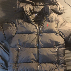  Polo Ralph Lauren jacka  - Säljer en svart dunjacka från Polo Ralph Lauren med huva och dragkedja. Har bara använt den i 3 veckor.Jackan har en broderad röd logotyp på bröstet och praktiska fickor med dragkedjor. Perfekt för kyliga dagar. Säljer den eftersom jag beställde fel storlek. Priset kan diskuteras 