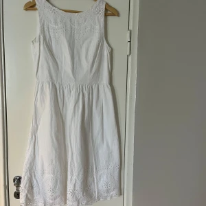 Vit ärmlös klänning med broderi - Charmig vit klänning med vackra broderade detaljer. Den är ärmlös och har en luftig passform, perfekt för varma dagar. Klänningen har ett fint mönster längs fållen och runt halsen som ger en romantisk touch.