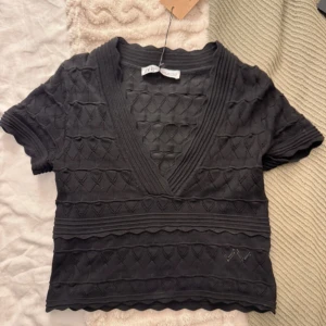 Svart v-ringad topp från Zara - Snygg svart topp från Zara med v-ringning och korta ärmar. Toppen har ett unikt stickat mönster och vågiga kanter som ger en trendig look. Perfekt för att lyfta vilken outfit som helst.