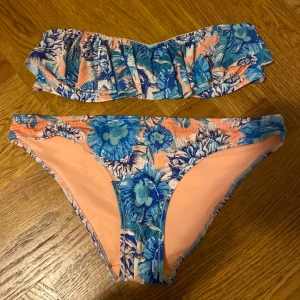 Blommig bikini från H&M - Snygg bikini från H&M med blommigt mönster i blått och orange. Överdelen är axelbandslös med volangdetaljer och underdelen har en klassisk passform. Perfekt för sommarens stranddagar!